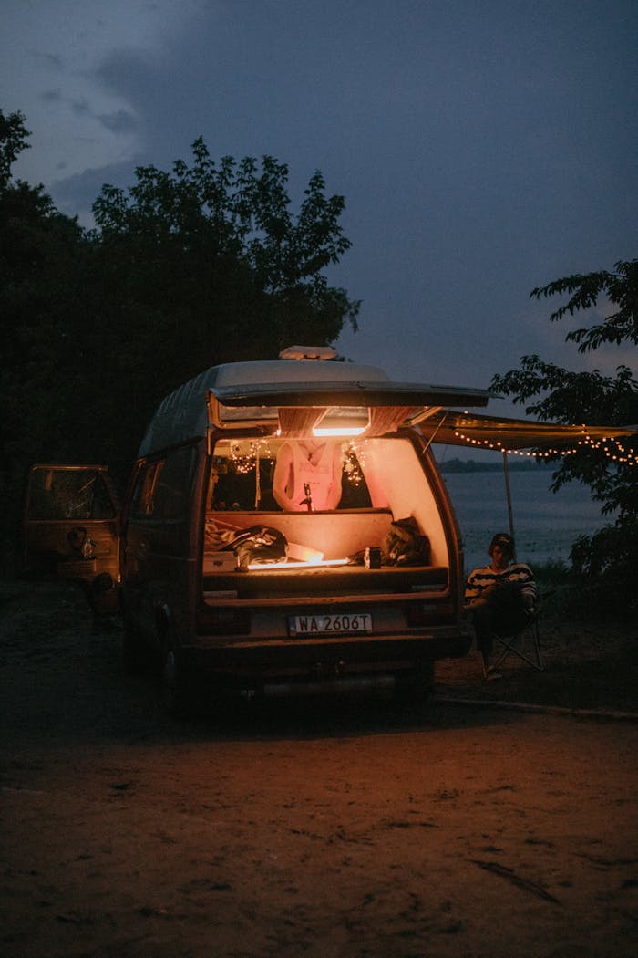 A person enjoys a peaceful evening by a lit camper van parked outdoors.