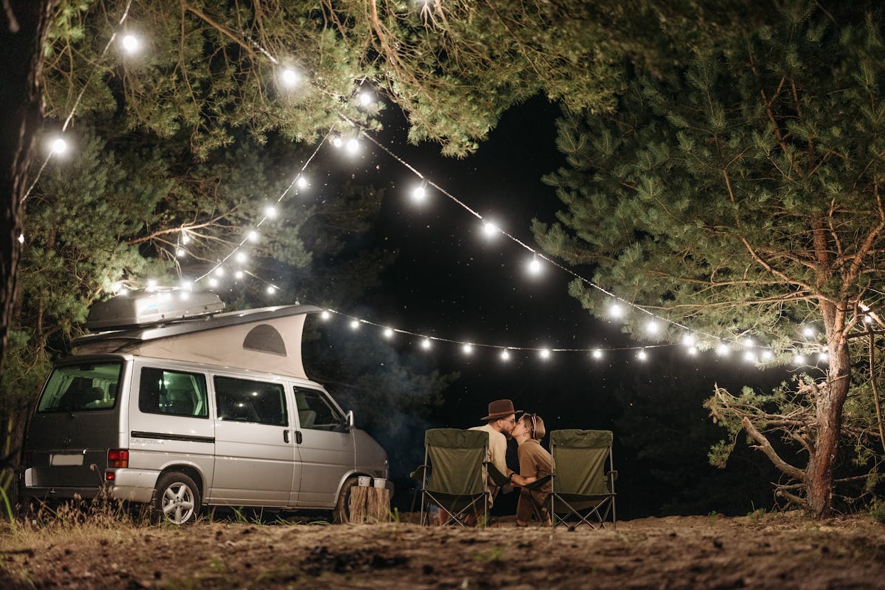 Couple sitting by a campervan enjoying a romantic night under string lights in a forest.